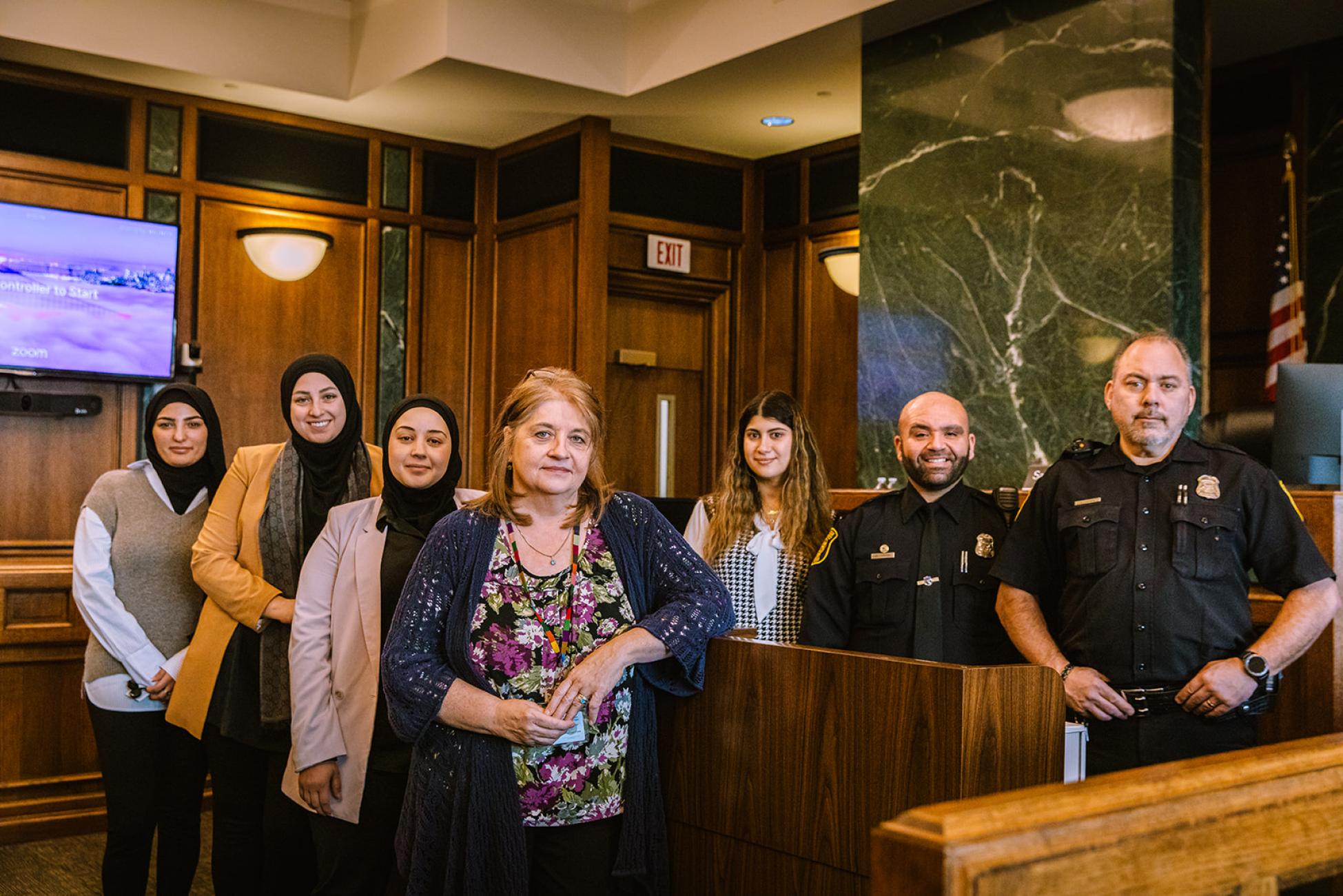 Seven members of the Court Staff pose for a photo inside the wood-paneled court chambers. 