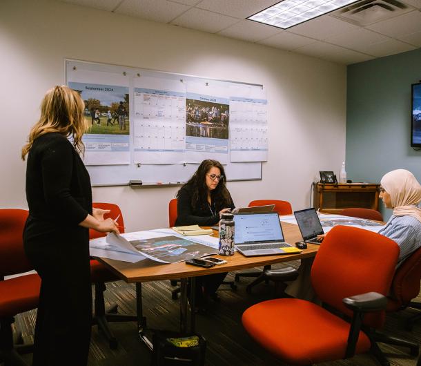 Three female members of the Communications Department work together around a conference table. A large print of the 2025 City of Dearborn Calendar is hanging up on the wall. 